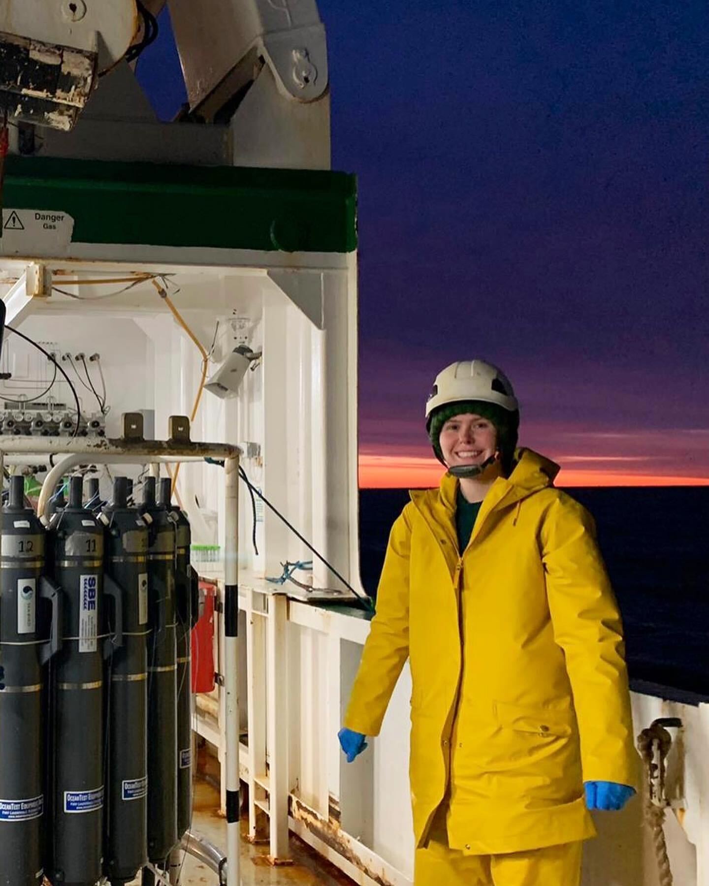 A student wearing a yellow coat and helmet stands on a ship's deck with the ocean and a sunset behind her